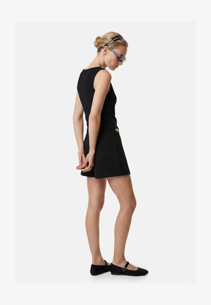 Frau trägt ein schwarzes ärmelloses Oberteil und einen Rock mit schwarzen Mary Jane Ballerinas, Sonnenbrille und einem doppelten schwarzen Haarband, steht und schaut nach unten.