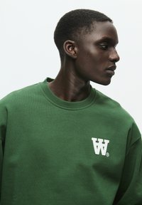 Green cotton sweatshirt featuring a round neckline and a white embroidered logo on the left chest. Smooth texture with a relaxed fit.