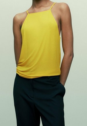 Persona che indossa una canottiera gialla senza maniche con spalline sottili e pantaloni neri, in piedi con una mano in tasca davanti a uno sfondo semplice.