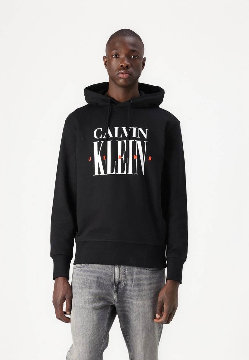 Juodos su gobtuvu džemperis su balta ir raudona "CALVIN KLEIN JEANS" grafika, ilgomis rankovėmis ir banguotu apvadu. Dėvimas su šviesiai pilkais džinsais.