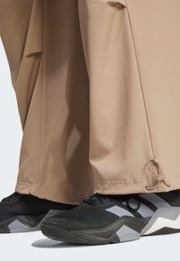 Beżowe spodnie o szerokich nogawkach z wiązaniem przy mankiecie, w połączeniu z czarnymi butami sportowymi z szarymi akcentami i teksturowanym materiałem.