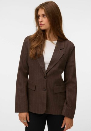 Femme aux longs cheveux bruns portant un blazer marron à fines rayures sur un haut blanc, regardant vers sa gauche sur un fond uni.