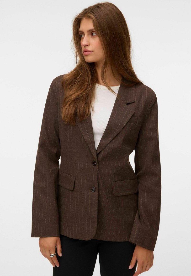 Femme aux longs cheveux bruns portant un blazer marron à fines rayures sur un haut blanc, regardant vers sa gauche sur un fond uni.