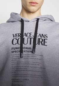 Šedá mikina s kapucí z měkké tkaniny s předním grafickým potiskem "VERSACE JEANS COUTURE" v tučném černém písmu a s šňůrkami na stažení.