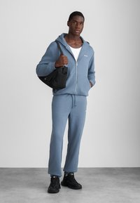 Hellblaue Zip-up Kapuzenjacke mit vorderen Taschen, kombiniert mit passenden Sweatpants. Das Model hält eine schwarze Tasche und trägt schwarze Sneakers.