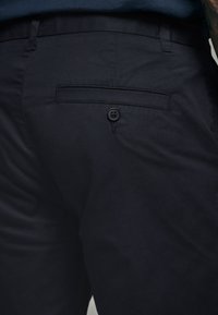 Dunkelblaue Hose aus Baumwollmischgewebe, ausgestattet mit einer Gesäßtasche mit einem einfachen Knopfverschluss und einer glatten Textur.