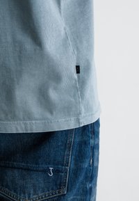 Lichtblauw, geribbeld katoenen T-shirt met een ronde zoom, voorzien van een klein zwart label bij de naad. Gecombineerd met donkerblauwe spijkerbroeken.