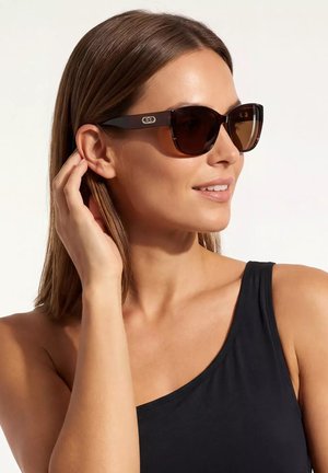 Kobieta nosząca ciemne, prostokątne okulary przeciwsłoneczne i czarny top z jednym ramieniem, dotykająca włosów, patrząca w bok na jednolitym tle.