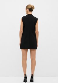 Vestido negro sin mangas con un cuello alto y cremallera en la parte posterior. Ajustado, de longitud por encima de la rodilla, de textura suave, combinado con zapatos de tacón alto negros.
