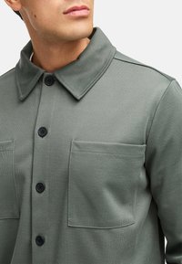 Skjorte med knaplukning i tekstureret, olivengrønt stof. Har en krave med spread collar, to brystlommer og sorte knapper til lukning.