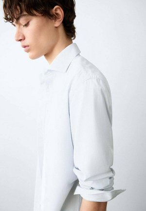 Giovane con capelli corti e ricci, che indossa una camicia chiara a maniche lunghe con le maniche arrotolate, guarda verso il basso su uno sfondo semplice.