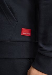 Felpa navy con tessuto testurizzato, dotata di un piccolo badge rettangolare rosso con la scritta "JOOP! JEANS" nella zona della tasca frontale.