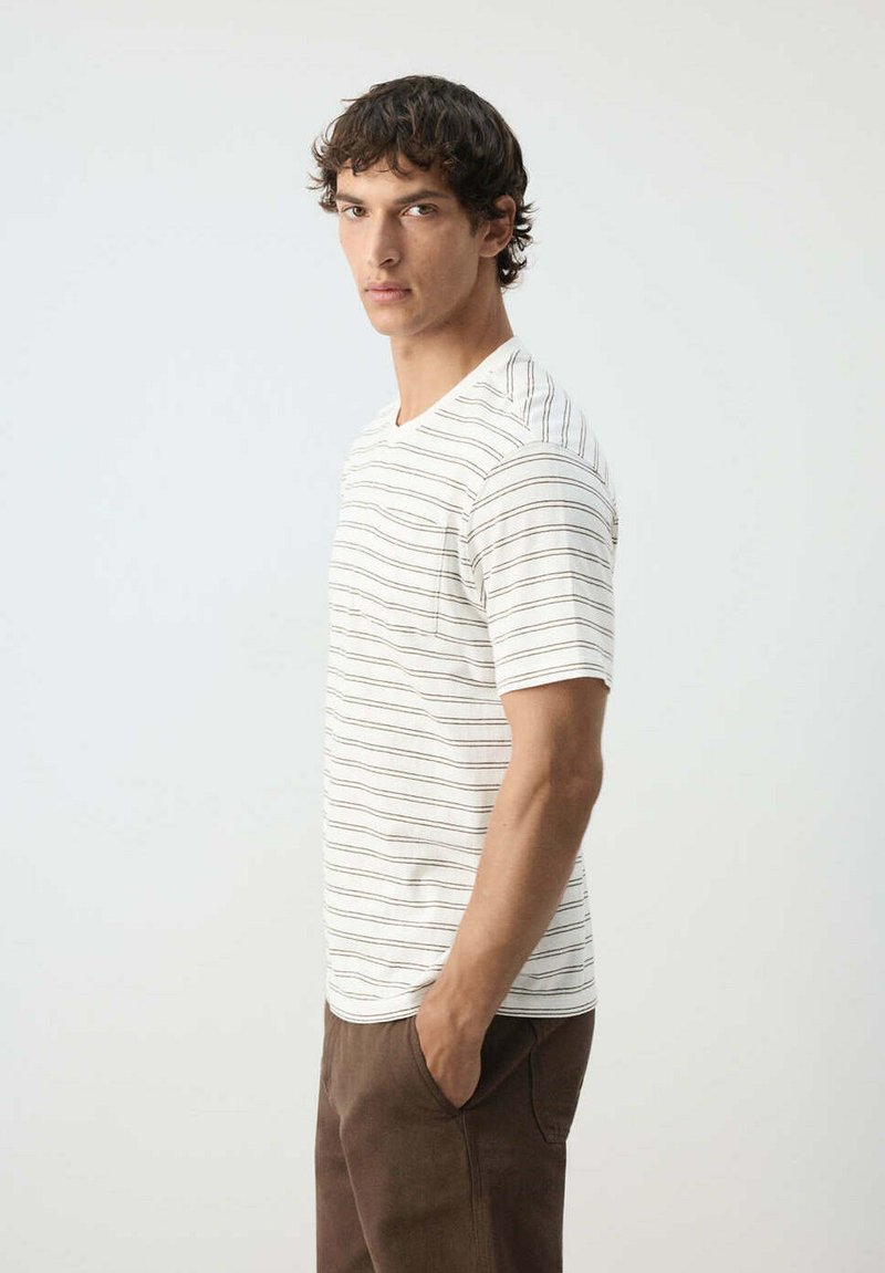 Joven de pie de lado, vistiendo una camisa blanca de manga corta con finas rayas horizontales y pantalones marrones, con una mano en el bolsillo.