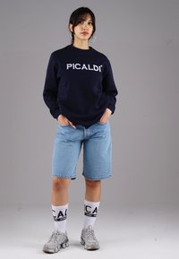 Navy sweatshirt with "PICALDI" logo, light blue denim shorts, white socks with black lettering, and silver athletic shoes on a model.