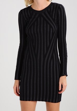 Femme portant une robe noire moulante à manches longues avec des rayures verticales et diagonales gris foncé.