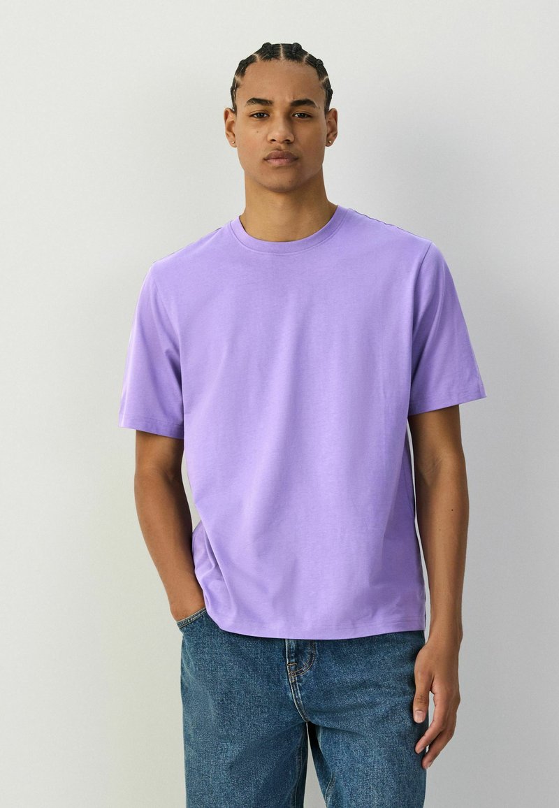 Jeune homme avec des tresses cornrow portant un t-shirt lavande uni et un jean bleu, debout devant un fond gris clair.