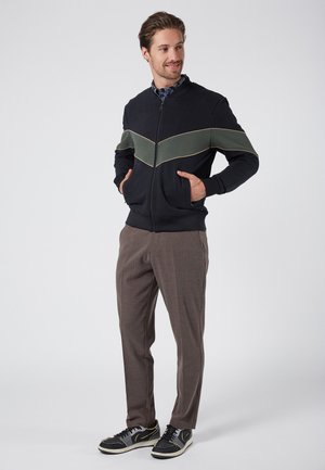 Black zip-up jacket with a green chevron stripe, paired with brown plaid trousers and black sneakers. Full front view, hands in pockets.