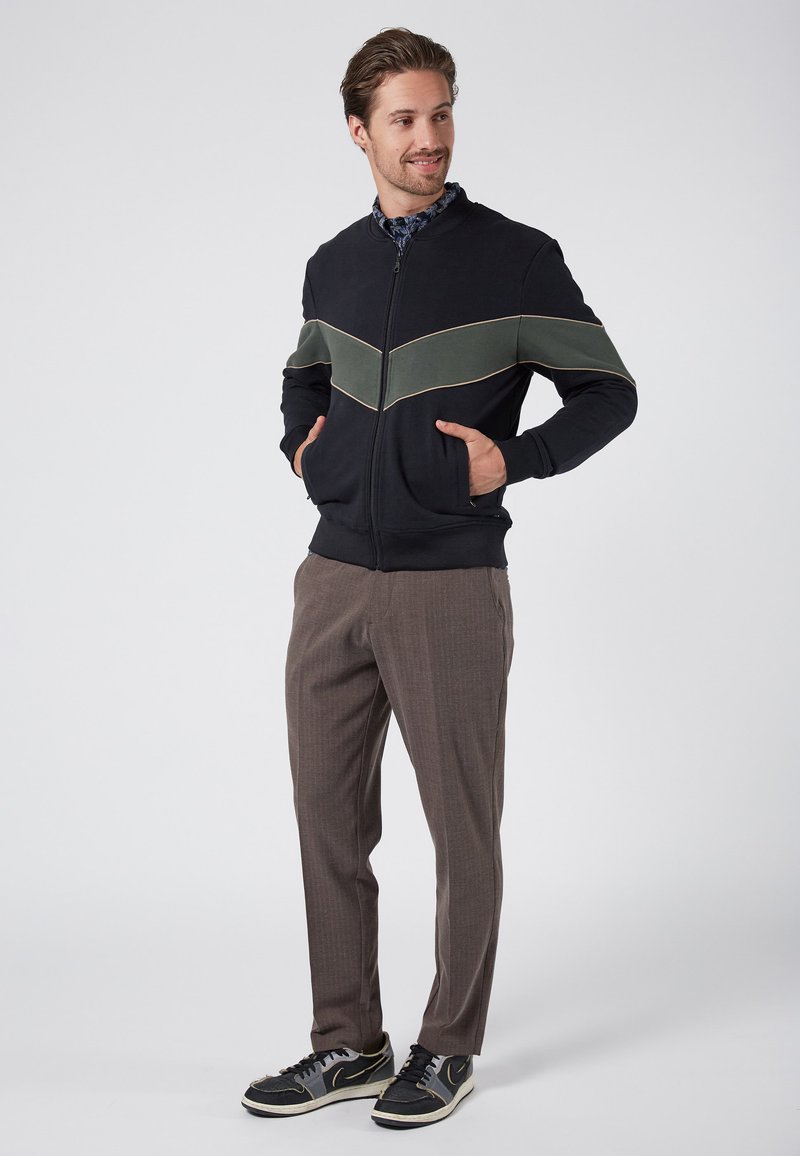 Giacca nera con zip, dotata di una striscia a forma di chevron verde, abbinata a pantaloni a quadri marroni e scarpe da ginnastica nere. Vista frontale completa, mani in tasca.