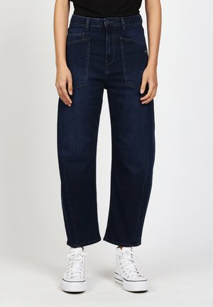 Woman wearing dark blue wide-leg jeans with front patch pockets and white high-top sneakers against a plain background.