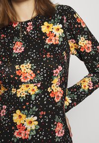 Nainen pitää yllä mustaa pitkähihaista mekkoa, jossa on keltaisia, oransseja ja vaaleanpunaisia kukkakuoseja sekä pieniä satunnaisia valkoisia ja punaisia kohtia.