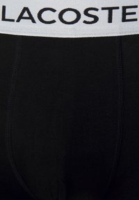 Black cotton boxer briefs with a white elastic waistband featuring "LACOSTE" in black lettering. Smooth texture and fitted design.