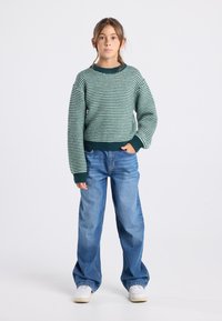 Maglione a righe verde e crema con scollatura rotonda, abbinato a jeans in denim blu e sneaker bianche, in piedi contro uno sfondo neutro.