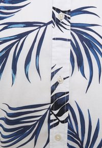 Biała tkanina z niebieskim i granatowym wzorem liści palmowych oraz trzema widocznymi białymi guzikami wzdłuż przeszytej listwy.