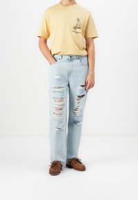Light yellow cotton t-shirt with a graphic print on the chest, paired with light blue ripped denim jeans featuring large distressed patches and fraying.