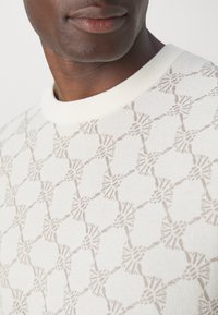 Gestrickter Pullover in hellem Creme mit einem strukturierten geometrischen Muster in Beige. Gerippter Kragen und glatte Stofftextur.