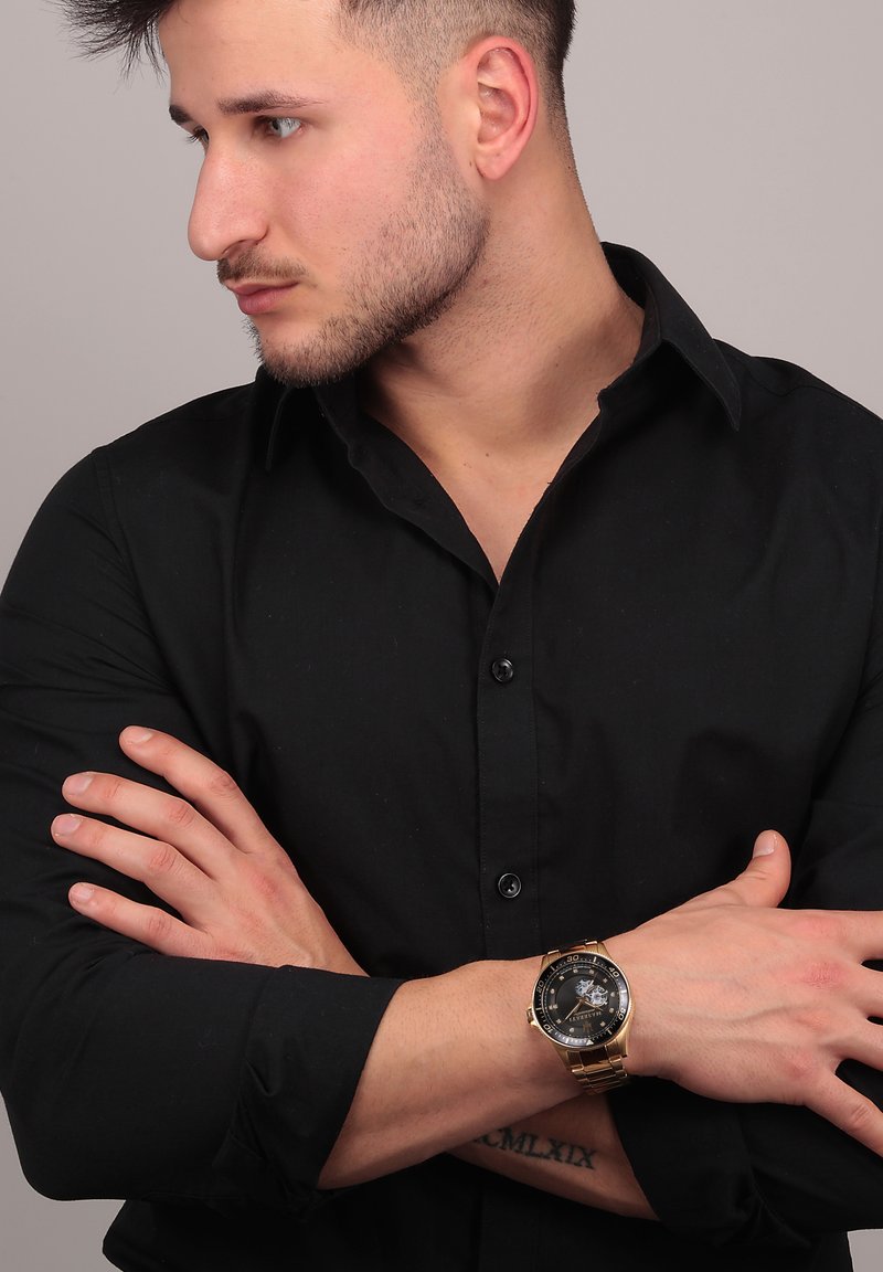 Guldtonet ur med sort urskive, metalarmbånd og detaljeret urskive, båret af en person i en tætsiddende sort skjorte med armene over kors.