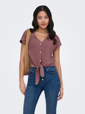 Femme portant un haut mauve à manches courtes avec boutons et noué à l'avant, un jean bleu taille haute, et un sac à bandoulière tissé sur l'épaule droite.
