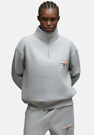 Jeune personne aux cheveux tressés portant un sweat zippé gris clair et un pantalon assorti, tous deux avec de petits accents orange.