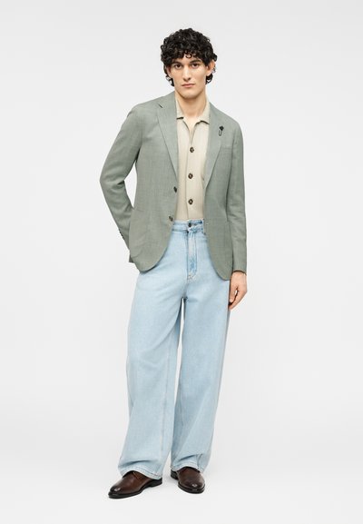 Ανοιχτό πράσινο σακάκι πάνω από μπεζ πουκάμισο με κουμπιά, συνδυασμένο με χαλαρά ανοιχτό μπλε τζιν και καφέ παπούτσια. Απλός σχεδιασμός, χωρίς ορατά σχέδια.