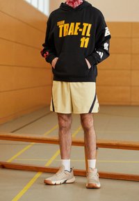 Sweat à capuche noir avec texte et graphismes jaunes, short jaune avec bandes latérales, et chaussures de sport beiges avec détails texturés.