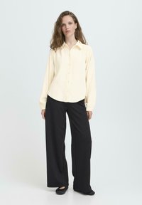 Camisa de botones crema de manga larga y corte relajado, combinada con pantalones negros de pierna ancha y zapatos planos negros sobre un fondo claro.