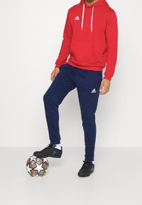Punainen huppari, jossa on valkoiset nyörit, tummansiniset verkkarit, joissa on Adidas-logo, sekä mustat jalkapallokengät jalkapallon päällä.