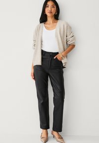 Cardigan beige oversize, top blanc, pantalon en cuir noir et talons couleur fauve. Le cardigan présente des manches amples ; le pantalon a une texture lisse avec une coupe droite.