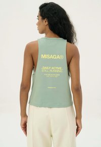 Mintgroene mouwloze tanktop met gele tekst. Heeft een ronde halslijn en open zijkanten. Het materiaal lijkt zacht en lichtgewicht, ideaal voor sportkleding.
