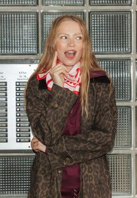 Manteau en imprimé léopard avec des tons marron foncé et beige, porté avec une écharpe à motifs rouge et blanc sur un haut bordeaux, devant un mur en briques de verre.