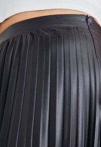 Gonna plissettata nera realizzata in materiale lucido. Presenta una cintura liscia e pieghe verticali, che mettono in evidenza la texture e il movimento.