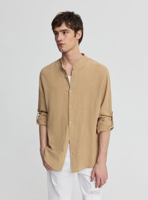 Młody mężczyzna z krótkimi brązowymi włosami, ubrany w beżową koszulę zapinaną na guziki z podwiniętymi rękawami oraz białe spodnie z rozdarciem na kolanie.