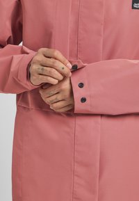 Roze waterdichte jas met een strak ontwerp, lange mouwen en zwarte knopen op de manchetten voor een veilige sluiting.