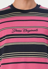 Dryžuota marškinėliai rožinės, tamsiai mėlynos ir grietinėlės spalvų; turi išsiuvinėtą "Guess Originals" logotipą; trumpomis rankovėmis ir apvaliu kaklu.