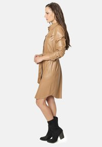Mujer con vestido de botones de cuero color beige y botas de tacón negras hasta el tobillo, de pie de perfil frente a un fondo blanco.