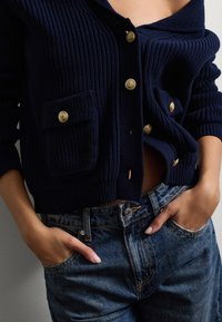 Marineblaues geripptes Strickjacke mit goldenen Knöpfen, ausgestattet mit zwei Fronttaschen und einem offenen Kragen. Kombiniert mit ausgebleichten blauen Jeans.