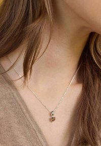 Collar de cadenas de plata con dos colgantes en forma de corazón, uno claro y otro ámbar, descansando sobre un suéter marrón claro.