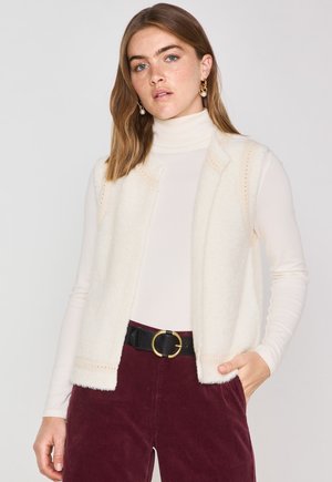 Gilet di lana color crema con una texture morbida, apertura frontale e cuciture decorative lungo i bordi, indossato sopra un dolcevita bianco aderente.