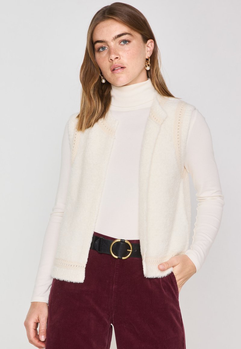 Gilet en laine de couleur crème avec une texture douce, à l'avant ouvert et avec une couture décorative le long des bords, porté sur un col roulé blanc ajusté.