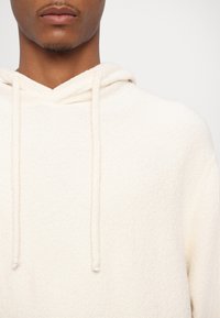 Gros plan d'une personne portant un sweat à capuche texturé de couleur crème avec des cordons de serrage, montrant la partie inférieure du visage et le cou.
