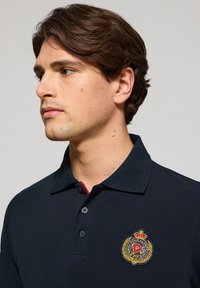 Polo de color azul marino con cuello acanalado, tapeta de tres botones y logo bordado que presenta un diseño de corona y laurel en el pecho.